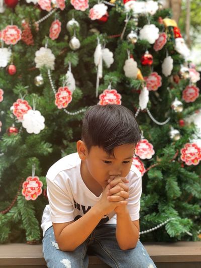 Boy looking away while sitting against christmas tree