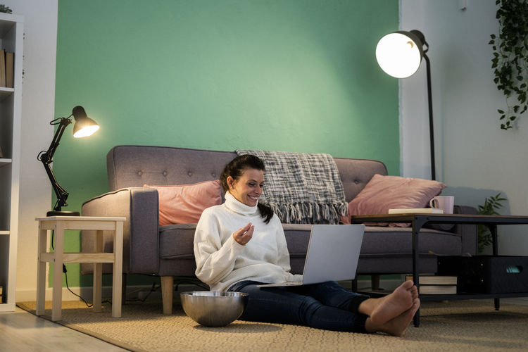 Mid adult woman using laptop while eating food sitting on floor at home