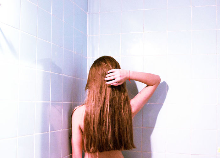 Rear view of shirtless woman with long brown hair against tiled wall