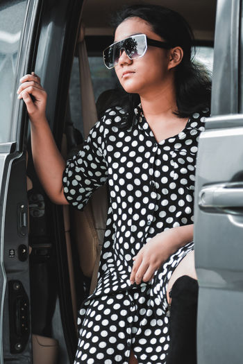 A girl wearing a polkadot dress, sun glasses and high boots sitting in a car 