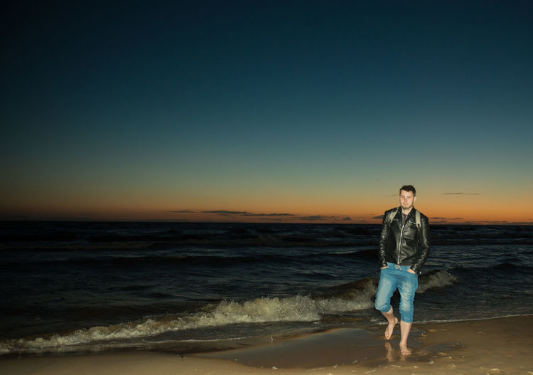Young man walking on shore at beach against sky during sunset
