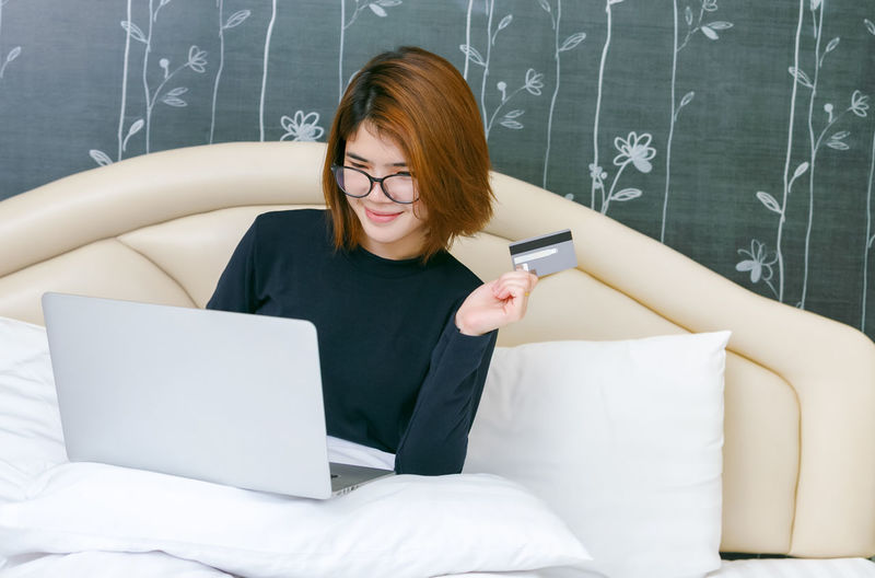 Smiling young woman doing online shopping over laptop while sitting on bed against wall at home