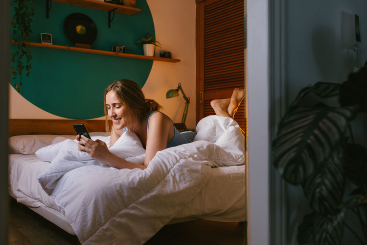 Smiley young woman texting with a friend in a modern bedroom.