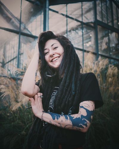 Smiling young woman with tattoo and dreadlocks against window 