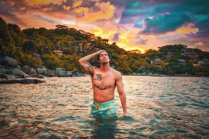 Portrait of shirtless man standing in mexican sea against sky during sunset