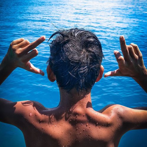 Rear view of shirtless teenage boy showing middle fingers in sea