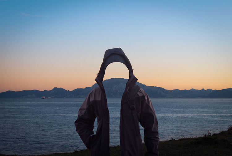 Digital composite image of invisible person wearing hooded jacket while standing by sea against clear sky during sunset