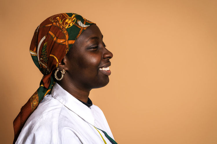 Cheerful african american female model in trendy headscarf and white shirt laughing with closed eyes on beige background in studio