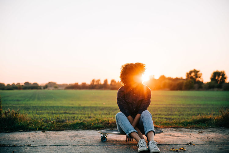 Teenage girl sitting on skateboard against clear sky during sunset