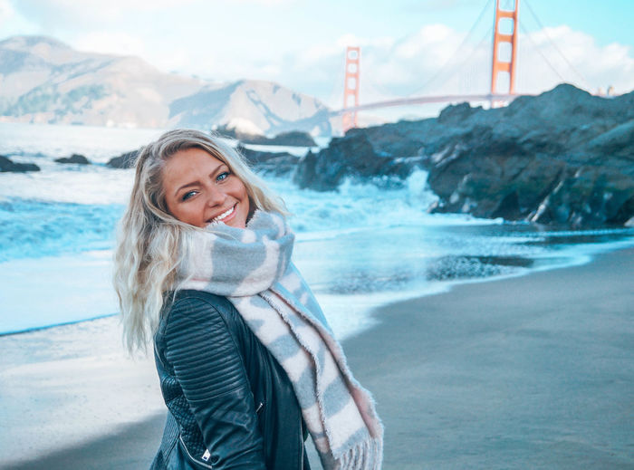 Portrait of smiling young woman standing at beach against golden gate bridge 