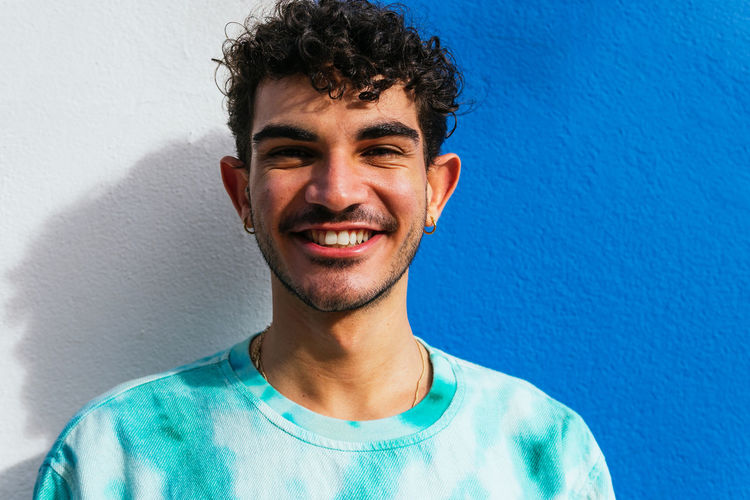 Happy young hispanic male in casual clothes leaning on two color wall and looking at camera with smile in daytime