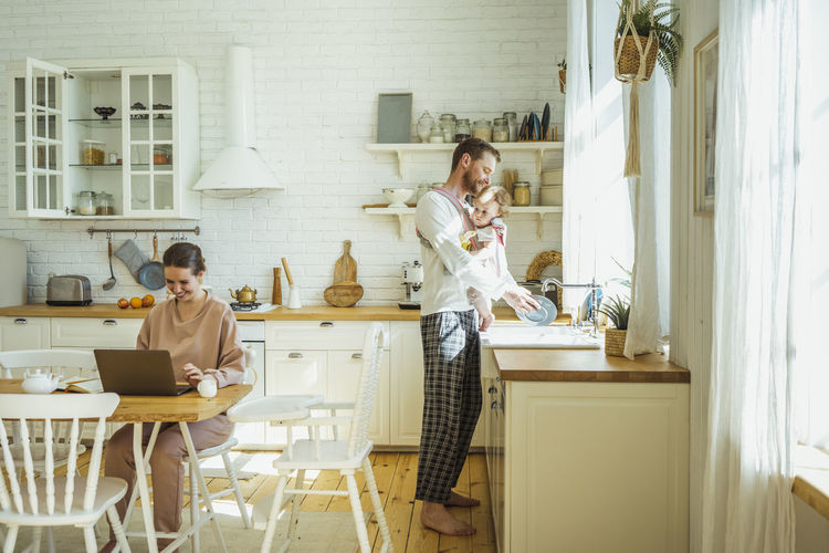 Smiling businesswoman using laptop and man carrying daughter while cleaning plate at kitchen