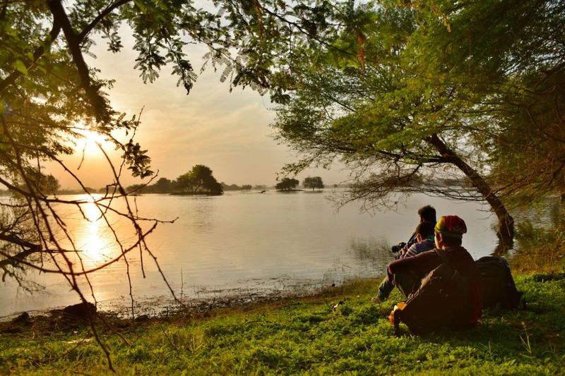 Friends sitting on grassy field by lake against sky during sunrise