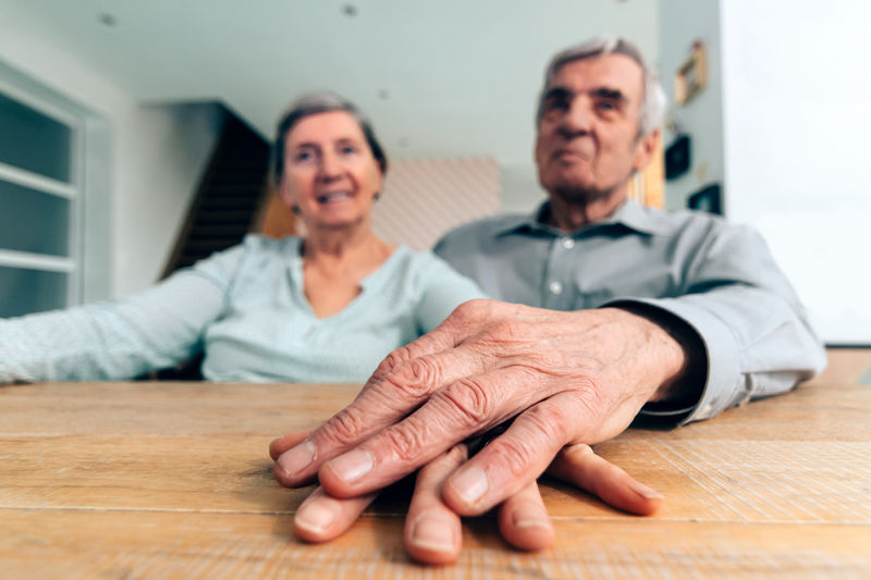 Elderly couple at home sitting at the table holding hand on hand - focus on hands in foreground