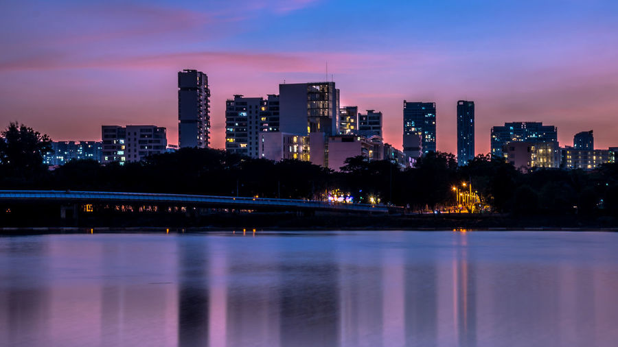 Illuminated buildings by river against sky at sunset
