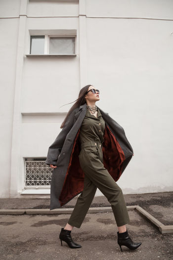 Full length side view postttait of young woman in grey coat walking against wall outdoors, outwear 