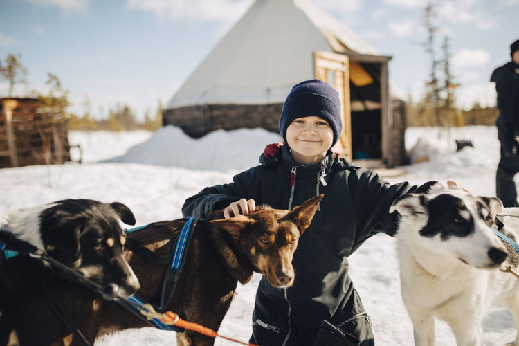 Portrait of boy wearing knit hat stroking husky dogs during winter