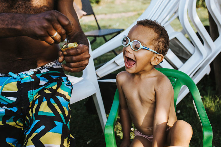 black father feeding his son a kiwi fruit Family Fatherhood Moments Feeding Fun Boy Child Childhood Emotion Enjoyment Father Focus On Foreground Fruit Glasses Innocence Kiwi Leisure Activity Lifestyles Mouth Open Outdoors Portrait Real People Shirtless Son Summer Two People Moms & Dads