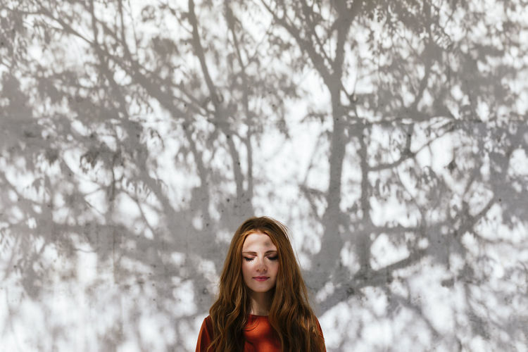 Redhead woman standing with eyes closed against tree shadow wall
