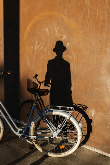 Shabby wall with shadow of unrecognizable male in hat near bicycle parked on paved sidewalk on sunny street of city
