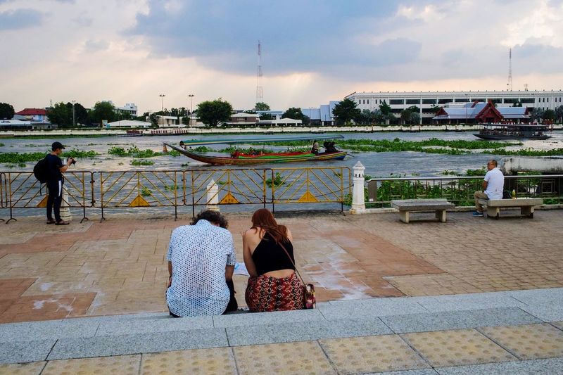 Women sitting on riverbank against sky in city