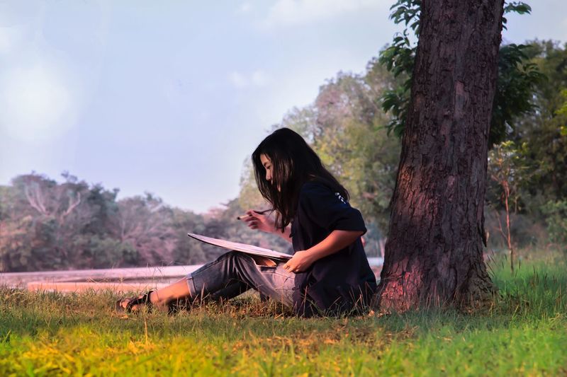 Side view of woman painting while sitting by tree trunk on field