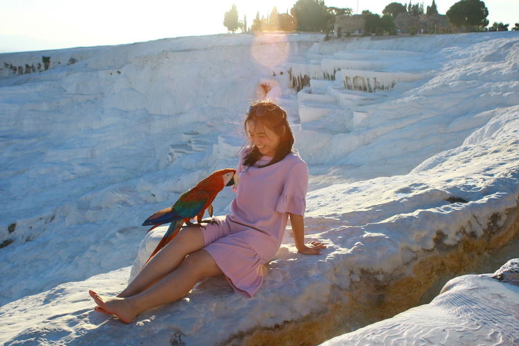 Full length of young woman sitting with bird on snow covered land during sunset