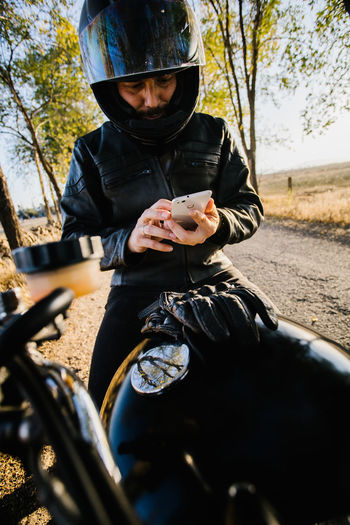 Concentrated male racer in leather jacket sitting on motorbike and browsing phone in autumn sunny day