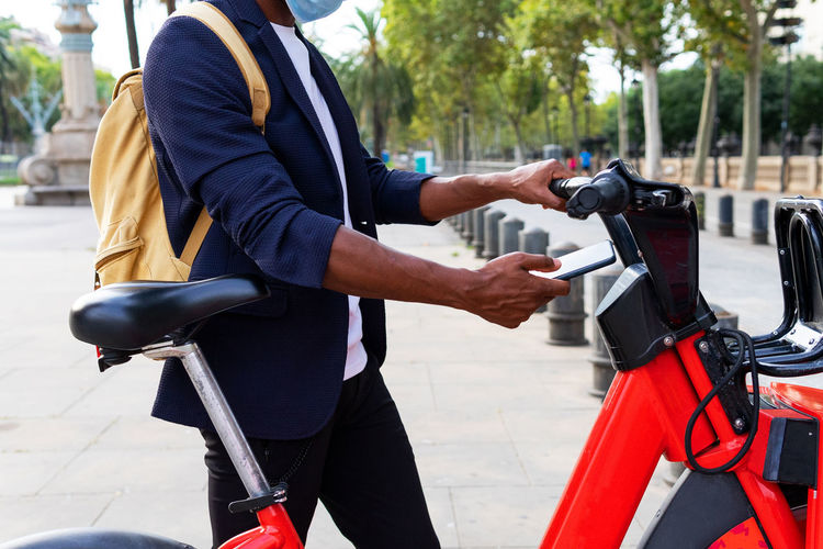 Black man in a suit and mask using his cell phone to rent a public bicycle on the street