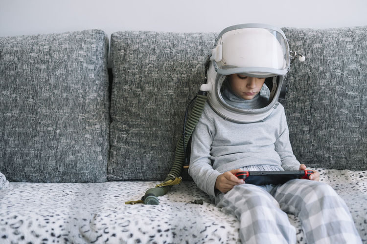 Adorable kid with a real astronaut uniform. he is sitting on couch and playing with a video game console at home