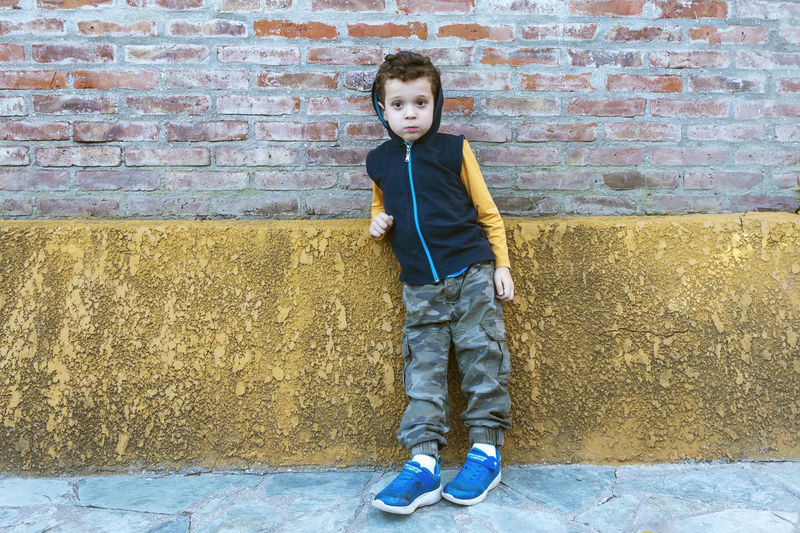 Boy in hooded sweatshirt and camouflage pants leaning against brick wall