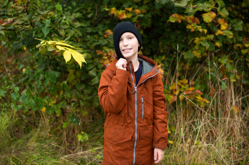 A cute boy holds a hat on his head with his hand near a yellow autumn tree