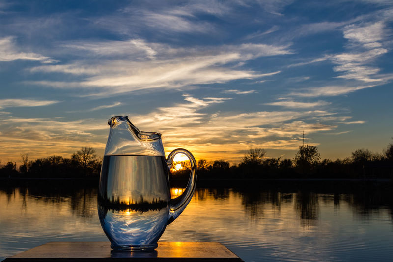 Lake seen through jug at table against sky during sunset
