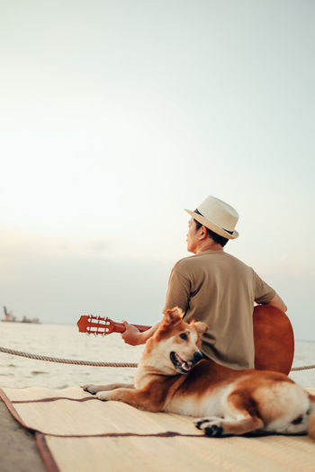 Rear view of man holding guitar sitting with dog against sky