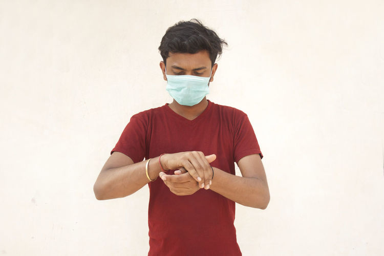 Young man wearing flu mask using hand sanitizer standing against white background