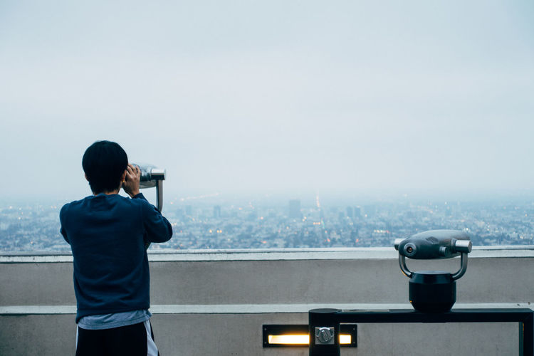 Rear view of man looking through binoculars against sky during foggy weather