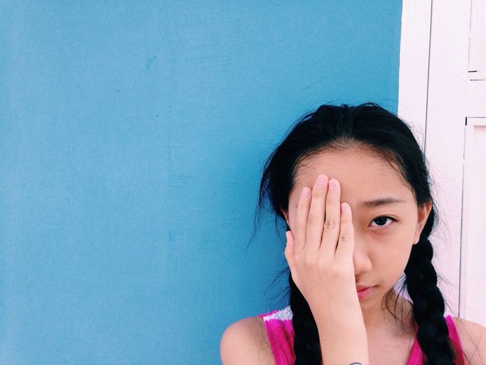 Portrait of girl covering eye with hand against blue wall