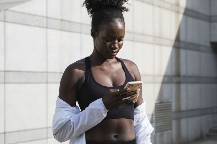 Slim african american female athlete standing on street on sunny day and messaging on mobile phone while having break during workout