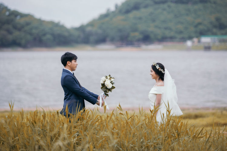 Married couple embracing amidst grass against lake