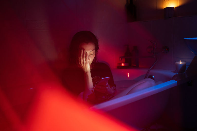 Unhappy woman with mental disorder, alcohol addiction sit in clothes in full bath holding smartphone