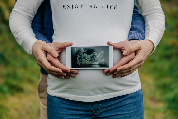 Midsection of pregnant couple holding mobile phone with ultrasound over abdomen while standing on grass 