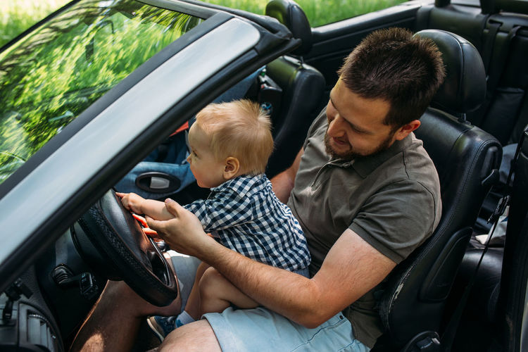 Dad shows his little son how to drive car while sitting behind wheel