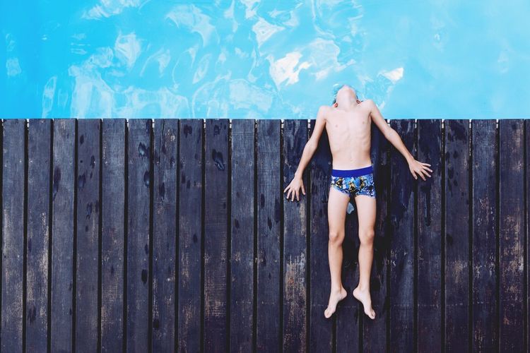 High angle view of boy lying on wooden deck by swimming pool