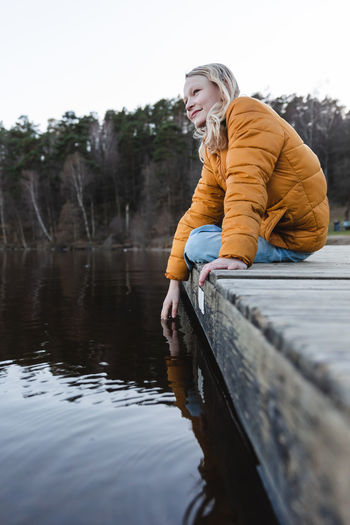 Low angle of cheerful teenage girl sitting on wooden quay near pond in autumn forest and looking away
