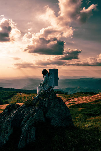 An astronaut sitting on rock against sky during sunset