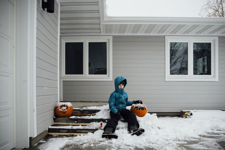 Portrait of girl in warm clothing sitting with halloween pumpkin on snow by house at backyard