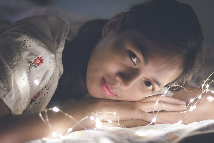 Close-up portrait of young woman lying by illuminated string lights on bed