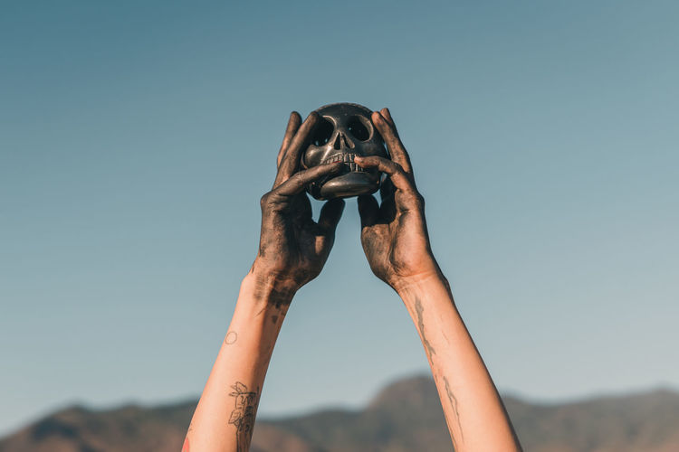 Female hands holding a black clay skull against clear blue sky in oaxaca mexico.
