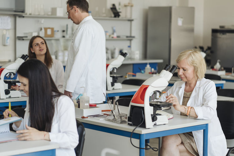 Senior female researcher in a white coat working in lab surrounded by other researchers