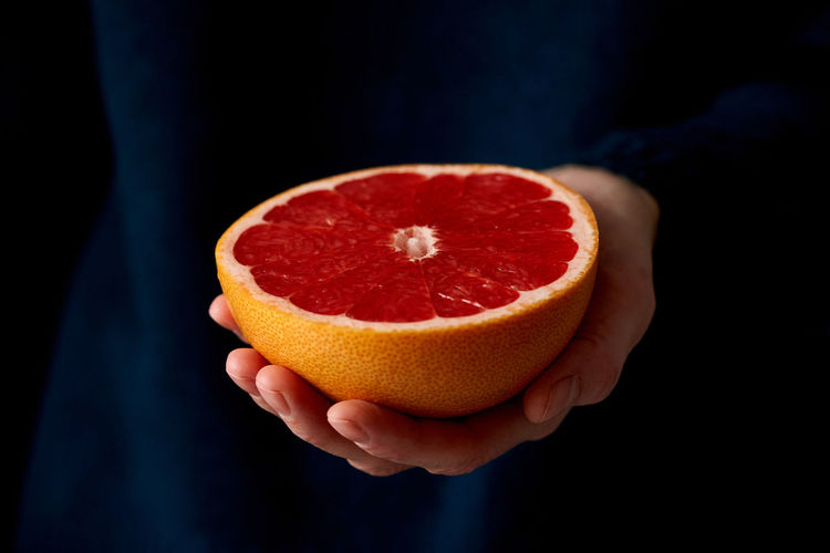 Close-up of woman's hand holding fresh halved red bright citrus fruit grapefruit on dark background